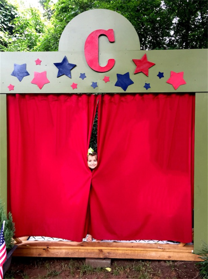 Green homemade puppet theater with red curtains, star cutouts and a child peeking through curtains