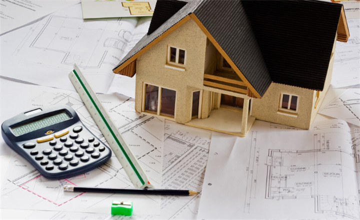 Miniature residential home model resting on site floor plans with calculator, ruler and drafting pencils for landscape and outdoor project costing